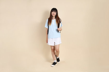 Asian woman wearing blue polo shirt,  white shorts, carrying school backpack, holding a book against a plain beige background. Gen Z,の写真素材