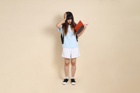 Asian woman wearing blue polo shirt,  white shorts, carrying school backpack, holding a book against a plain beige background. Gen Z,の写真素材