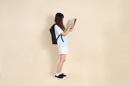 Asian woman wearing blue polo shirt,  white shorts, carrying school backpack, holding a book against a plain beige background. Gen Z,の写真素材