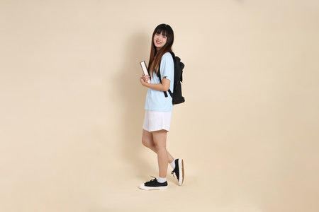 Asian woman wearing blue polo shirt,  white shorts, carrying school backpack, holding a book against a plain beige background. Gen Z,の写真素材