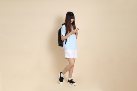 Asian woman wearing blue polo shirt,  white shorts, carrying school backpack, Holding mobile phone against a plain beige background. Gen Z,の写真素材