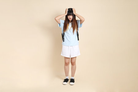 Asian woman wearing blue polo shirt,  white shorts, carrying school backpack, holding a book against a plain beige background. Gen Z,の写真素材