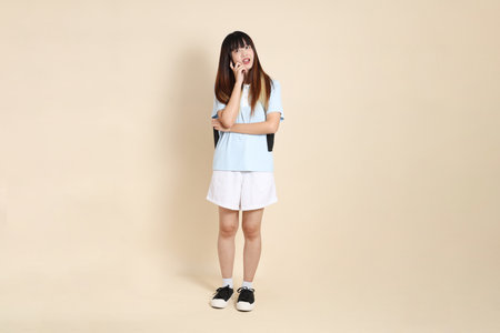 Asian woman wearing blue polo shirt,  white shorts, carrying school backpack, holding a book against a plain beige background. Gen Z,の写真素材