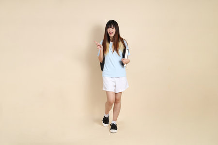 Asian woman wearing blue polo shirt,  white shorts, carrying school backpack, holding a book against a plain beige background. Gen Z,の写真素材