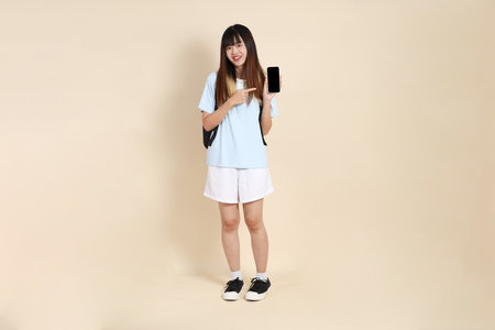 Asian woman wearing blue polo shirt,  white shorts, carrying school backpack, Holding mobile phone against a plain beige background. Gen Z,の写真素材