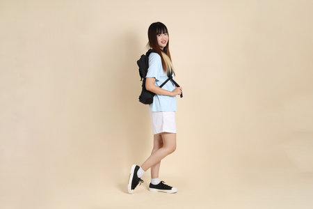 Asian woman wearing blue polo shirt,  white shorts, carrying school backpack, holding a book against a plain beige background. Gen Z,の写真素材