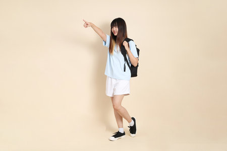Asian woman wearing blue polo shirt,  white shorts, carrying school backpack, holding a book against a plain beige background. Gen Z,の写真素材