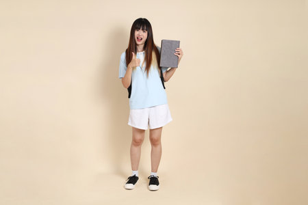 Asian woman wearing blue polo shirt,  white shorts, carrying school backpack, holding a book against a plain beige background. Gen Z,の写真素材