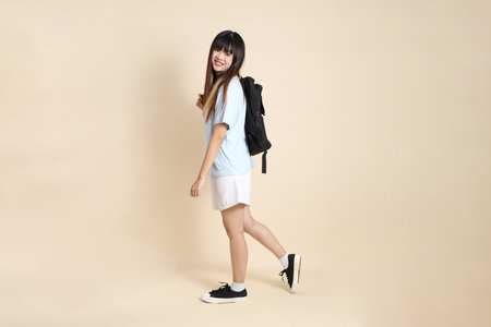 Asian woman wearing blue polo shirt,  white shorts, carrying school backpack, holding a book against a plain beige background. Gen Z,の写真素材