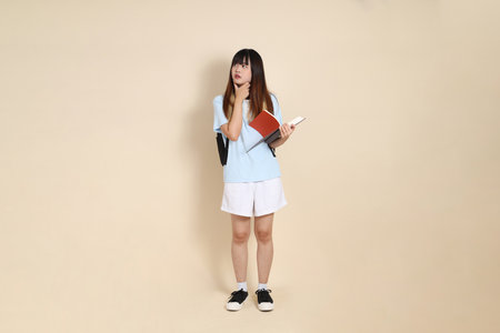 Asian woman wearing blue polo shirt,  white shorts, carrying school backpack, holding a book against a plain beige background. Gen Z,の写真素材