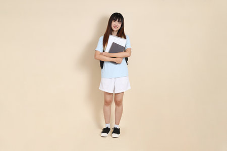 Asian woman wearing blue polo shirt,  white shorts, carrying school backpack, holding a book against a plain beige background. Gen Z,の写真素材