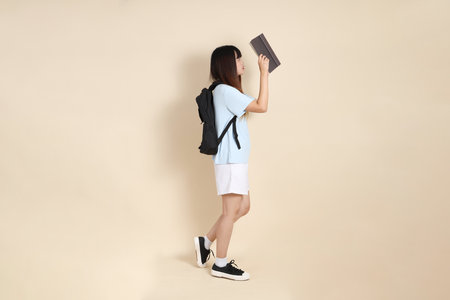 Asian woman wearing blue polo shirt,  white shorts, carrying school backpack, holding a book against a plain beige background. Gen Z,の写真素材