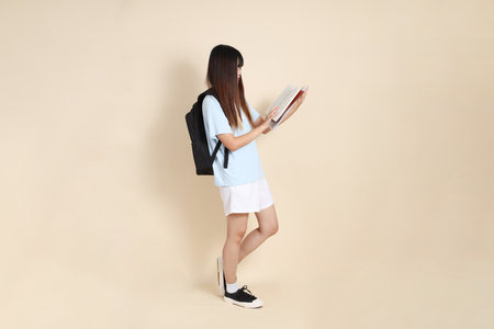 Asian woman wearing blue polo shirt,  white shorts, carrying school backpack, holding a book against a plain beige background. Gen Z,の写真素材