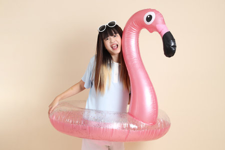 Smiling young Asian woman wearing sunglasses on head, holding a large pink flamingo inflatable swim ring against beige background. Summer holiday, vacation and lifestyle, College student, Gen Zの写真素材
