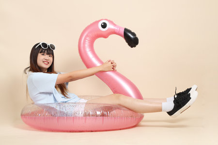 Smiling young Asian woman wearing sunglasses on head, holding a large pink flamingo inflatable swim ring against beige background. Summer holiday, vacation and lifestyle, College student, Gen Zの写真素材