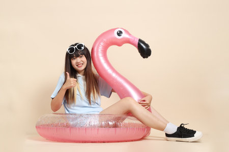 Smiling young Asian woman wearing sunglasses on head, holding a large pink flamingo inflatable swim ring against beige background. Summer holiday, vacation and lifestyle, College student, Gen Zの写真素材
