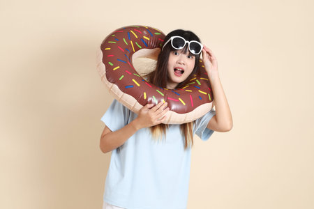 Smiling young Asian woman wearing sunglasses on head, holding an inflatable swim ring against beige...