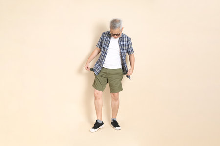 Portrait of an Asian senior man in glasses, empty pocket, wearing casual plaid shirt, shorts, and sneakers, isolated on beige background. Elderly male lifestyle concept.の写真素材