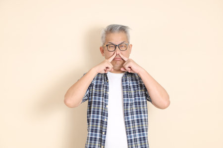 Portrait of an Asian senior man in glasses, cover nose, wearing casual plaid shirt, shorts, and sneakers, isolated on beige background. Elderly male lifestyle concept.の写真素材