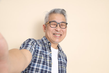 Portrait of an Asian senior man in glasses, taking a selfie, Holding a camera to taking a photo, wearing casual plaid shirt, shorts, and sneakers, isolated on beige background. Elderly male lifestyle concept.の写真素材