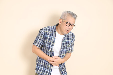 Portrait of an Asian senior man in glasses, stomachache, Abdominal pain, wearing casual plaid shirt, shorts, and sneakers, isolated on beige background. Elderly male lifestyle concept.の写真素材