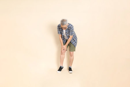 Portrait of an Asian senior man in glasses, wearing casual plaid shirt, shorts, and sneakers, isolated on beige background. Elderly male lifestyle concept.の写真素材