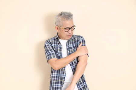 Portrait of an Asian senior man in glasses, arm ache, wearing casual plaid shirt, shorts, and sneakers, isolated on beige background. Elderly male lifestyle concept.の写真素材