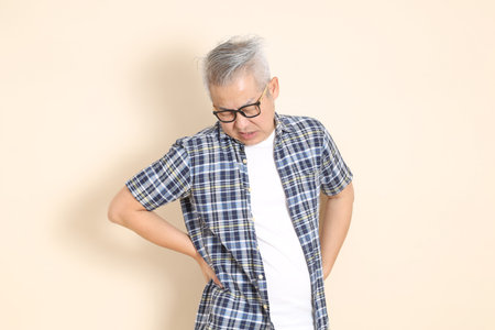 Portrait of an Asian senior man in glasses, wearing casual plaid shirt, shorts, and sneakers, isolated on beige background. Elderly male lifestyle concept.の写真素材