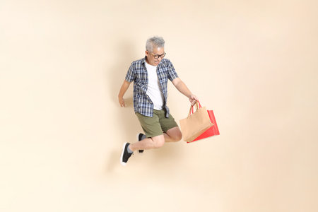 Portrait of an Asian senior man in glasses, holding shopping bag while jumping, wearing casual plaid shirt, shorts, and sneakers, isolated on beige background. Elderly male lifestyle concept.の写真素材