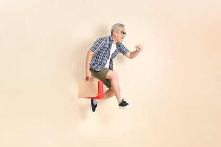 Portrait of an Asian senior man in glasses, holding shopping bags while jumping, wearing casual plaid shirt, shorts, and sneakers, isolated on beige background. Elderly male lifestyle concept.の写真素材