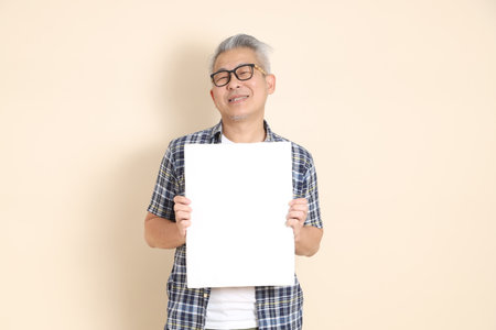 Portrait of an Asian senior man in glasses, holding blank canvas, wearing casual plaid shirt, shorts, and sneakers, isolated on beige background.の写真素材