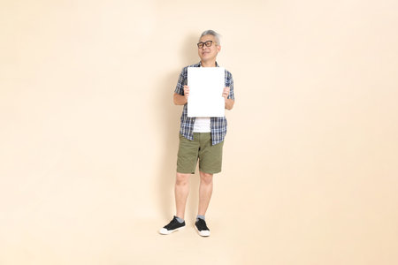 Portrait of an Asian senior man in glasses, Holding blank canvas, wearing casual plaid shirt, shorts, and sneakers, isolated on beige background. Elderly male lifestyle concept.の写真素材