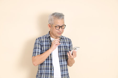 Portrait of an Asian senior man in glasses, Holding mobile phone, wearing casual plaid shirt, shorts, and sneakers, isolated on beige background. Elderly male lifestyle concept.の写真素材