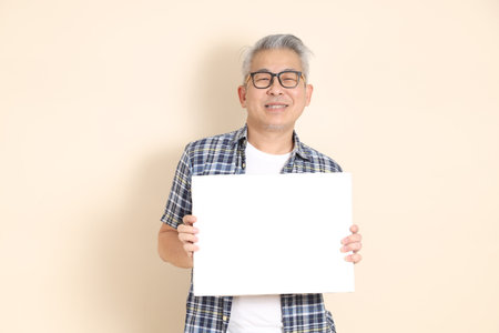 Portrait of an Asian senior man in glasses, Holding blank canvas, wearing casual plaid shirt, shorts, and sneakers, isolated on beige background. Elderly male lifestyle concept.の写真素材