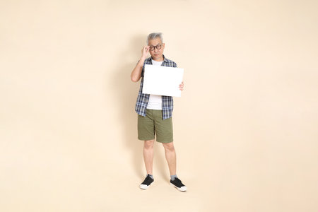 Portrait of an Asian senior man in glasses, Holding blank canvas, wearing casual plaid shirt, shorts, and sneakers, isolated on beige background. Elderly male lifestyle concept.の写真素材