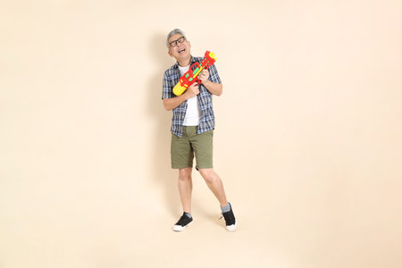 Portrait of an Asian senior man in glasses, holding a water gun, wearing a casual plaid shirt, shorts, and sneakers, isolated on a beige background. Elderly male lifestyle concept.の写真素材