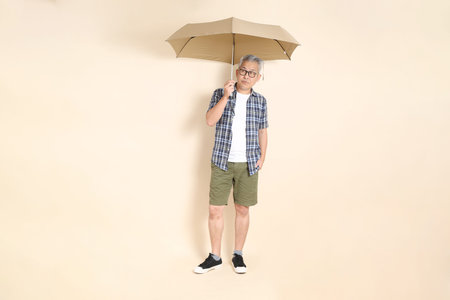 Portrait of an Asian senior man in glasses, Holding Umbrella, wearing casual plaid shirt, shorts, and sneakers, isolated on beige background. Elderly male lifestyle concept.の写真素材