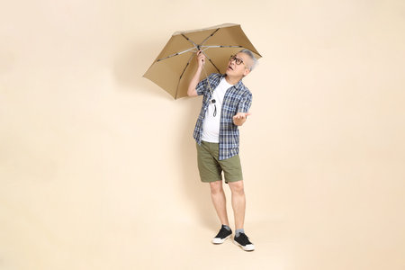 Portrait of an Asian senior man in glasses, Holding Umbrella, wearing casual plaid shirt, shorts, and sneakers, isolated on beige background. Elderly male lifestyle concept.の写真素材