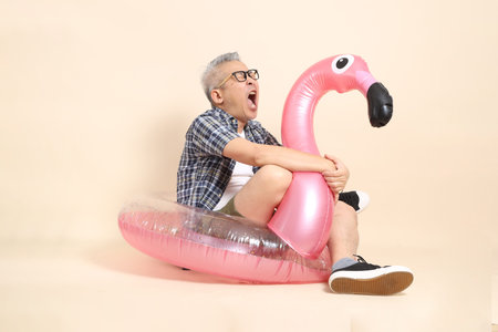 Asian senior man in glasses, holding a large pink flamingo inflatable swim ring , wearing casual plaid shirt isolated on beige background. Elderly male lifestyle concept.の写真素材