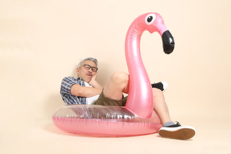 Asian senior man in glasses, holding a large pink flamingo inflatable swim ring , wearing casual plaid shirt isolated on beige background. Elderly male lifestyle concept.の写真素材