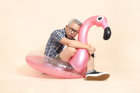 Asian senior man in glasses, holding a large pink flamingo inflatable swim ring , wearing casual...