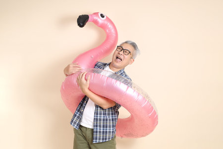 Asian senior man in glasses, holding a large pink flamingo inflatable swim ring , wearing casual plaid shirt isolated on beige background. Elderly male lifestyle concept.の写真素材