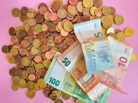 A stack of various coins alongside euro bills is displayed prominently on a soft pink surface, creating an eyecatching contrastの写真素材
