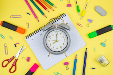 A variety of colorful stationery items creatively arranged alongside a stylish clock on a bright yellow backgroundの写真素材