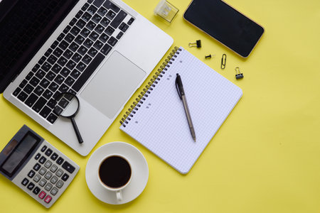 A vibrant office setup with a laptop, a warm coffee, and stationery against a cheerful yellow backdropの写真素材