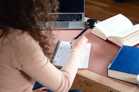 A focused student writing notes with a laptop and books atop a desk in a cozy, creative study...