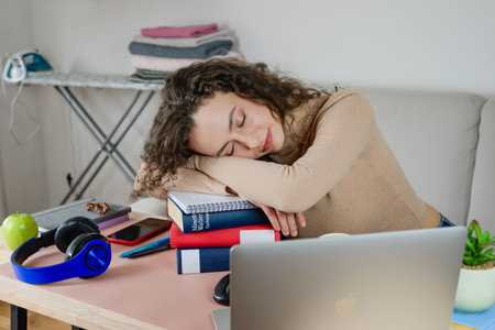 A tired student is diligently studying with multiple books and a laptop in their home space