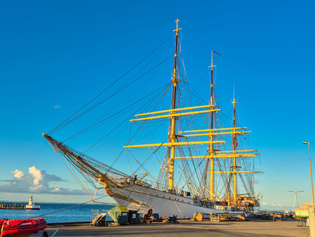 A magnificent tall ship majestically resting at the dock beneath a vibrant blue sky dotted with...