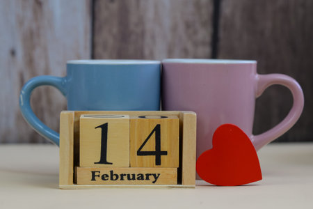 valentine's morning with two pastel mugs, wooden block calendar displaying 14 February, red heart cutout leaning against calendar,の写真素材