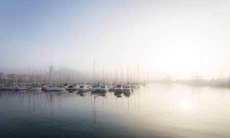 Photography of quiet and beautiful sea port, where stands many small and large yachts with masts, boats. The sky is gray and smooth and is a light fog.の写真素材